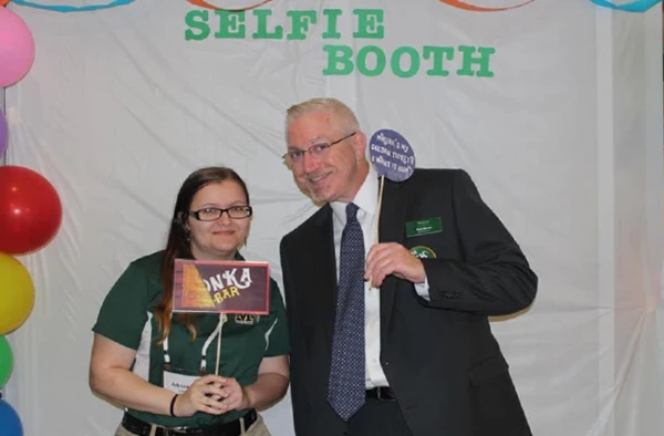 A woman and a man posing in front of a 'SELFIE BOOTH' backdrop with party decorations, the woman holding a book titled 'ANKA' and the man holding a speech bubble sign.